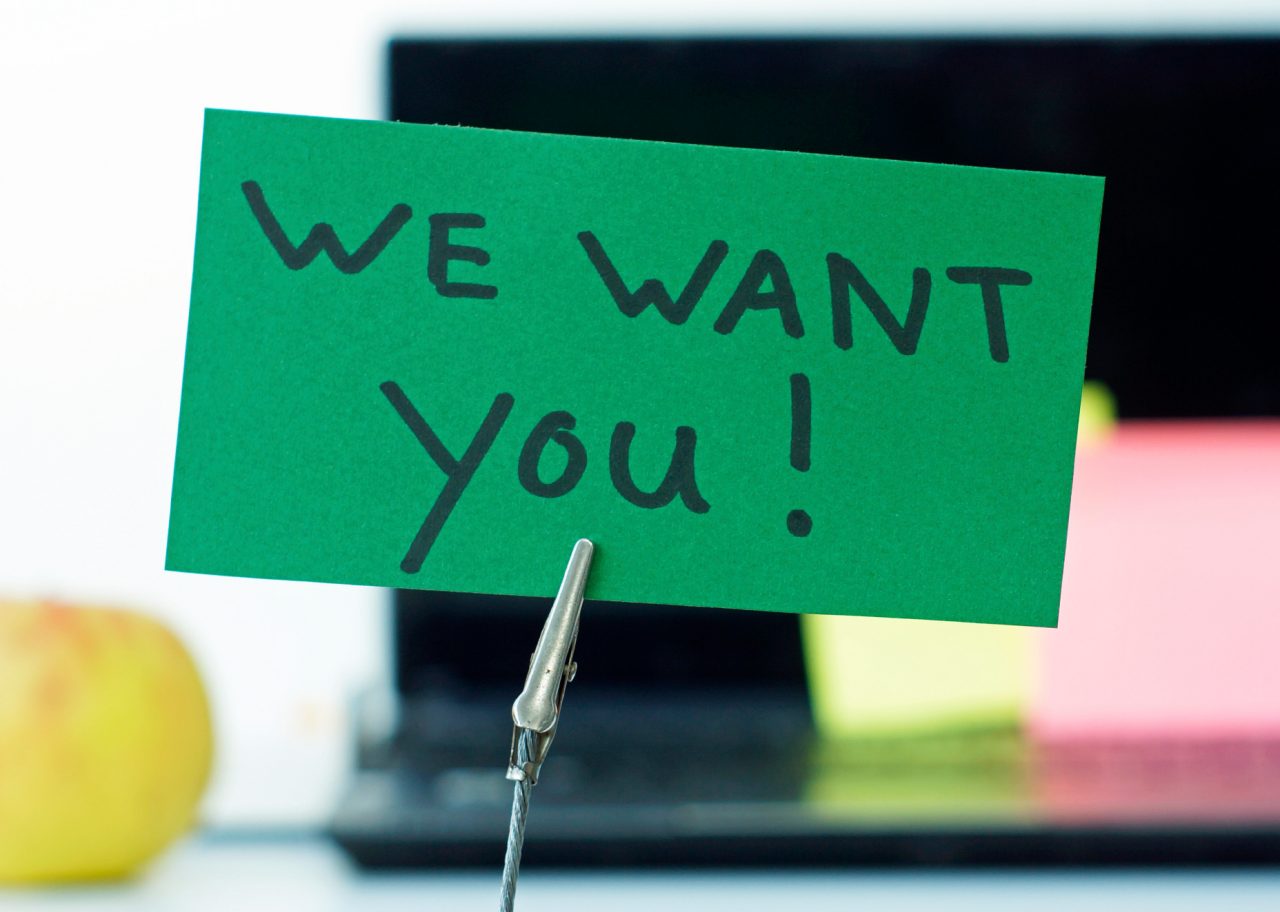 Green sign on desk that says "we want you!"