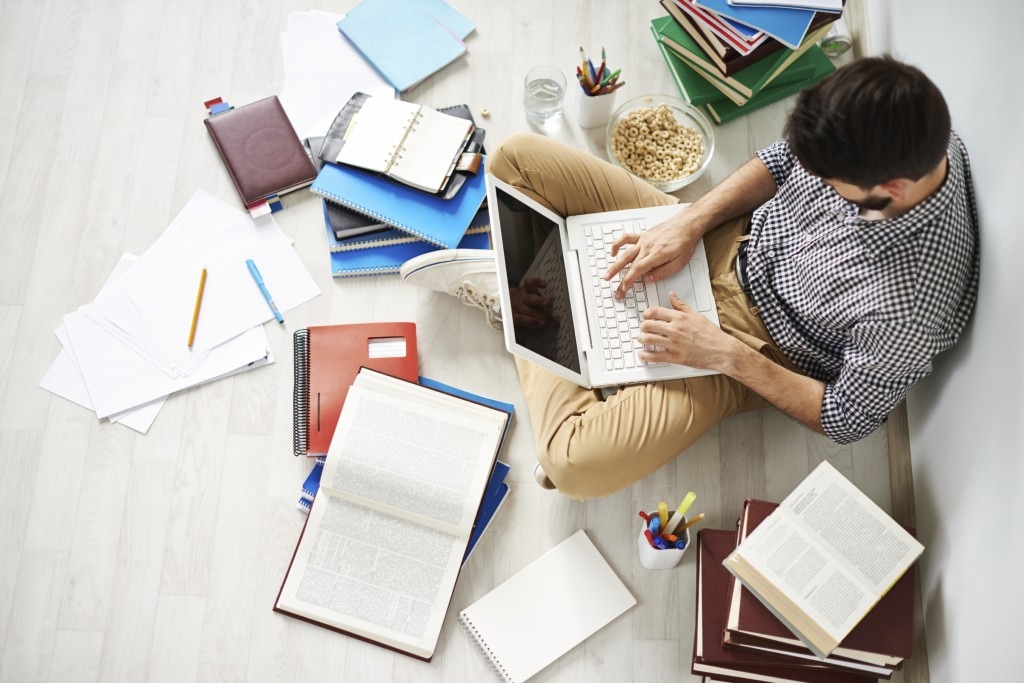 Étudiant assis par terre avec un ordinateur portable sur ses genoux entouré de cahiers, de livres et de papiers