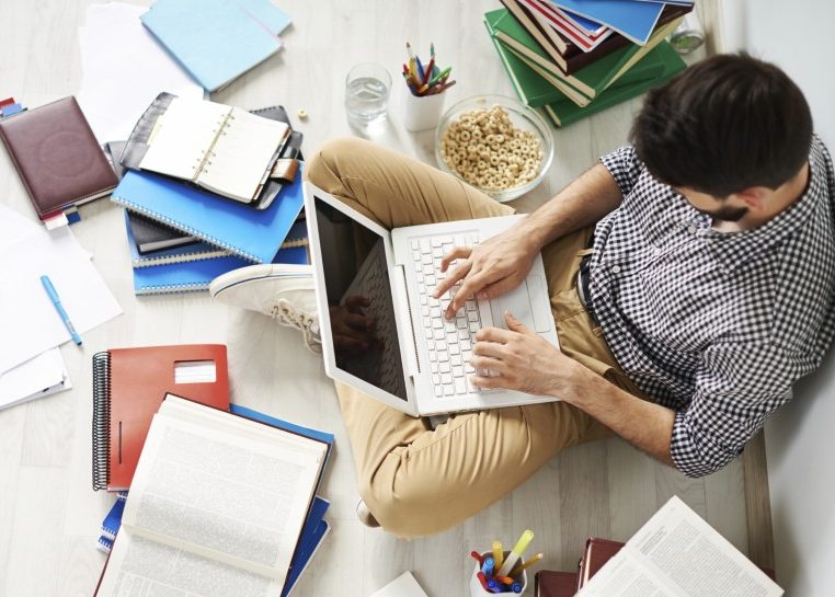 Étudiant assis par terre avec un ordinateur portable sur ses genoux entouré de cahiers, de livres et de papiers