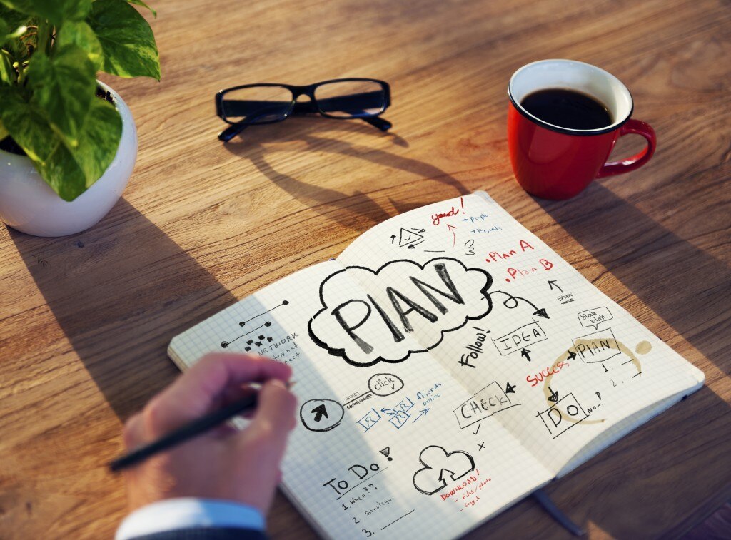 Écriture à la main dans un cahier avec des plans sur un bureau avec une plante, des verres et une tasse de café