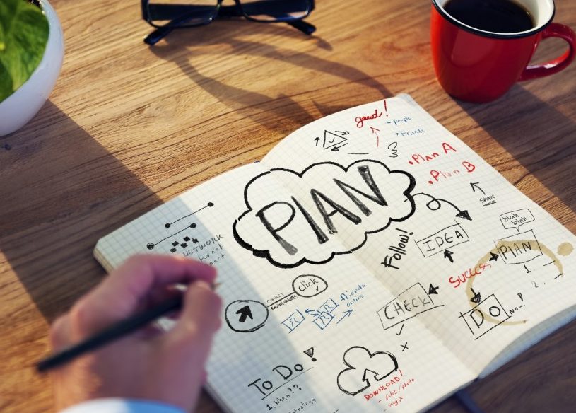 Écriture à la main dans un cahier avec des plans sur un bureau avec une plante, des verres et une tasse de café